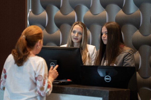 Two ladies standing behind a hotel check-in desk speak with a lady who is in front of the desk. 