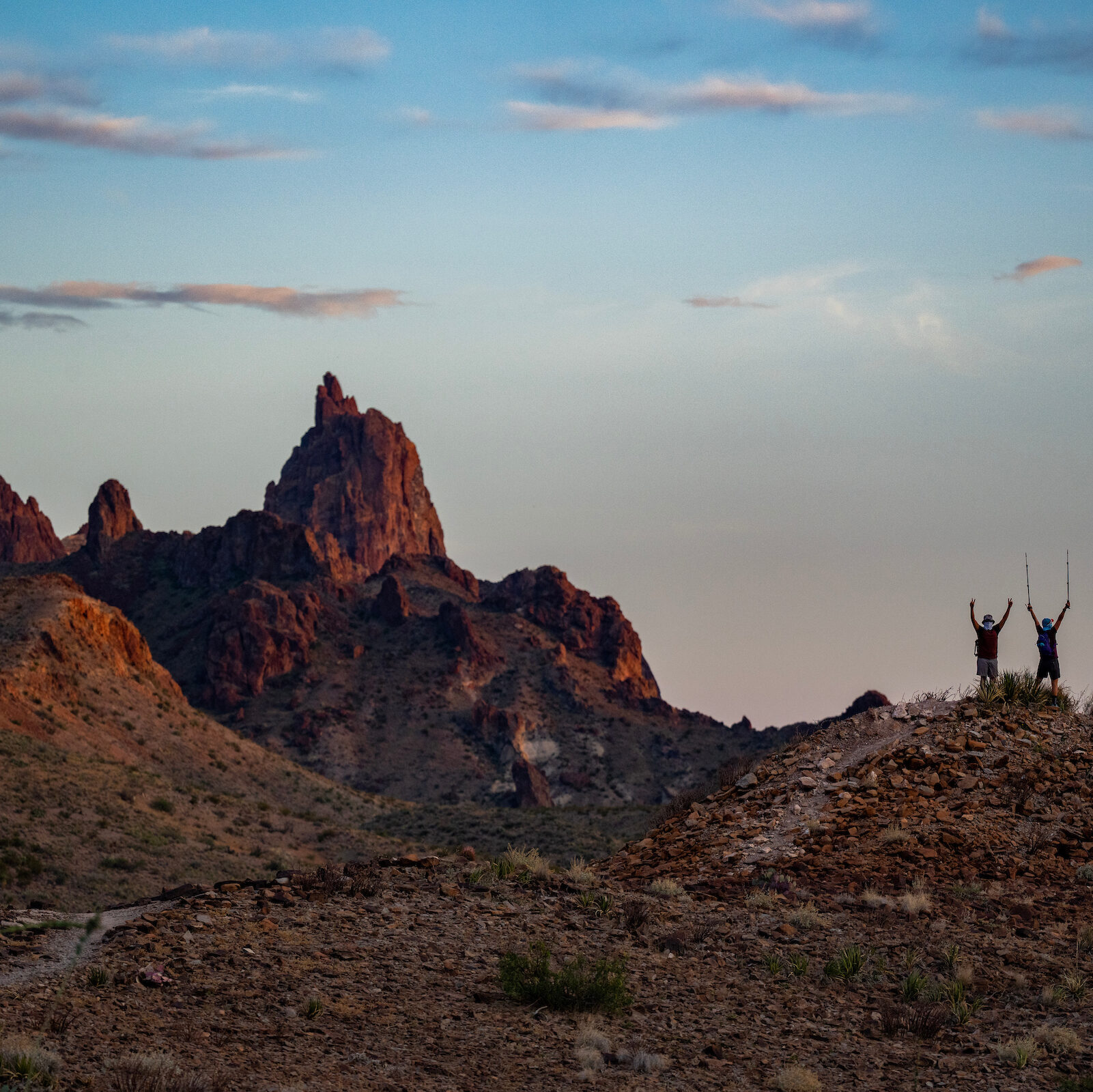 Rocky terrain with two people standing on a hill with their arms raised in the distance.